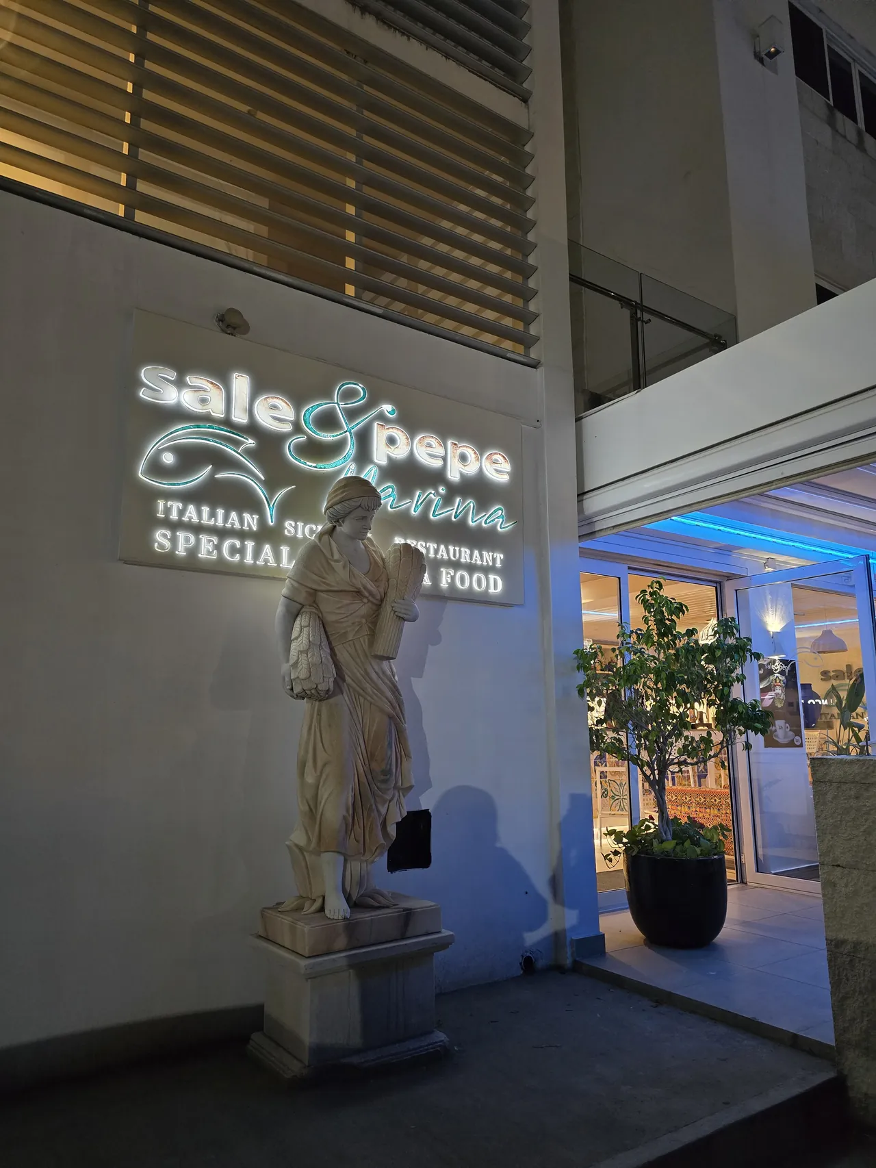 Entrée de Sale & Pepe avec statue siciliene en pierre devant l'enseigne lumineuse